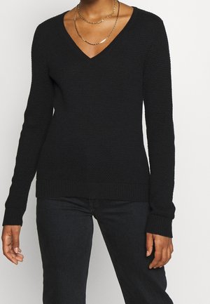 Persoon die een zwarte, gestructureerde V-hals trui, zwarte broek en twee gelaagde gouden kettingen draagt, staand tegen een effen achtergrond.