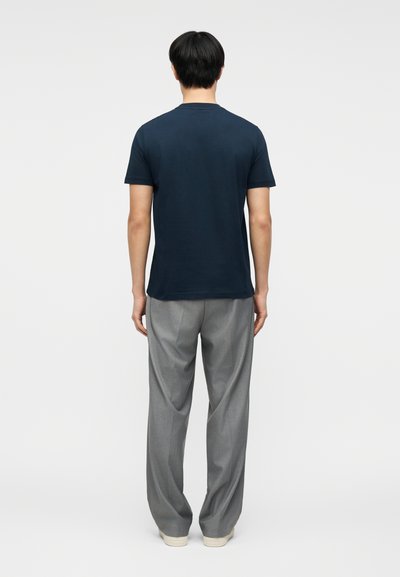 Άνδρας που στέκεται πλάτη φοράει μπλε σκούρο κοντομάνικο πουκάμισο, γκρι παντελόνι και λευκά παπούτσια μπροστά σε απλό φόντο.