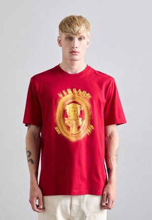 Blond mees, kes kannab punast Versace T-särki, millel on kollane Medusa embleem ja tekst, seistes ühetaolise heledashall tausta vastu.
