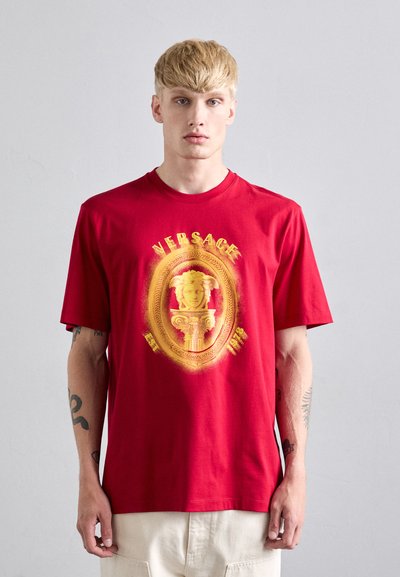 Blond mężczyzna w czerwonej koszulce Versace z żółtym emblematem Meduzy i napisem, stojący na jednolitym jasnoszarym tle.