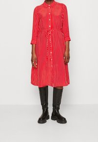 Abito rosso a button-up con strisce bianche verticali, maniche di lunghezza media, vita con cintura e gonna svasata. Indossato con stivali neri fino al ginocchio.