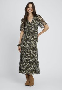 Robe maxi avec un décolleté en V, des manches courtes évasées et un design à volants en imprimé floral vert et noir. Associée à des bottines brunes.