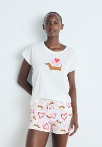Camiseta blanca de manga corta con un gráfico de un dachshund y globos de corazón coloridos. Combinada con pantalones cortos ligeros con un patrón de corazones y dachshunds.