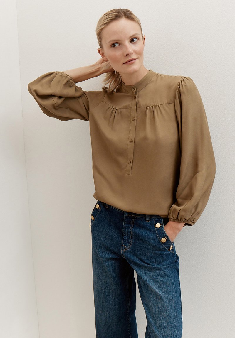 Blouse marron à col avec manches bouffantes, patte de boutonnage sur le devant et détails froncés, associée à un jean bleu avec des accents dorés.