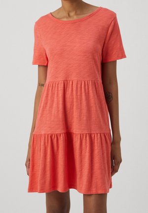 Kobieta ubrana w koralową sukienkę z krótkim rękawem, z falbanami i okrągłym dekoltem na jednolitym jasnym tle.