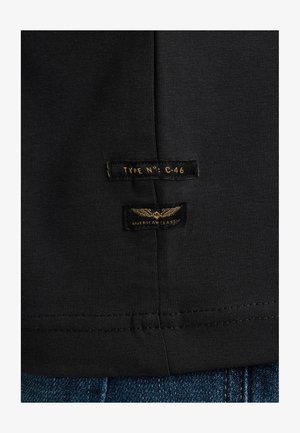 Sort bomulds t-shirt med to labels: "TYPE N°: C-46" og "AMERICAN CLASSIC," med guld tekst og vinge grafik. Syet kantdetalje.