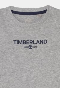 Šedé tričko s kulatým výstřihem, s námořnickou modrou logem Timberland a znakem stromu umístěným v centru pod výstřihem, velikost 8/126 označení uvnitř límce.
