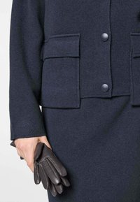 Marineblauwe gebreide stof met een textuur, voorzien van twee grote zakken, knoopsluiting en een hand die donkere leren handschoenen vasthoudt.