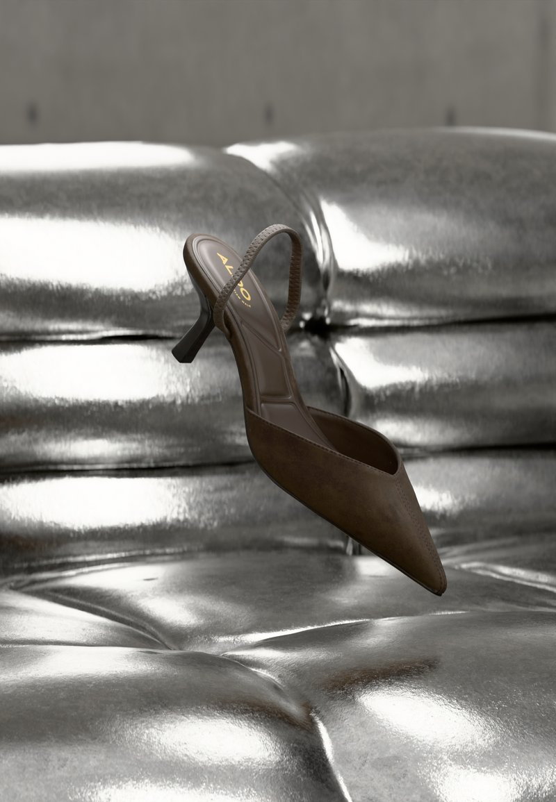Chaussure marron à bout pointu avec une fine bride, en suède, dotée d’un petit talon carré. Placée sur une surface métallique brillante.