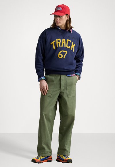 Sweat-shirt marine avec un graphique jaune "TRACK 67", un pantalon cargo vert, des baskets multicolores et une casquette rouge. Col en tissu visible.
