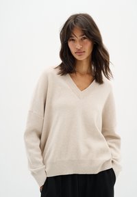 Pull en maille beige avec un col en V, texture douce, épaules tombantes et ourlet côtelé. Porté sur un pantalon noir sur fond uni.