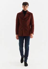 Brązowy dwurzędowy płaszcz marynarski wykonany z wełny, z dużymi czarnymi guzikami, kołnierzem z wycięciem i prostym krojem. Nosi się go z niebieskimi dżinsami.