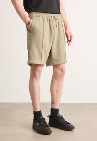 Light olive green shorts in textured fabric with an elastic waistband and drawstring. Features side pockets and a relaxed fit. Black sneakers.