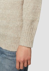 Beige gebreide trui met een gestructureerde afwerking en geribbelde manchetten, gecombineerd met lichtblauwe denim. De stof lijkt zacht en warm te zijn.