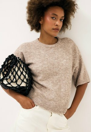 Mujer con cabello rizado que lleva un suéter de punto beige de manga corta y pantalones blancos, sosteniendo una bolsa de red negra con interior blanco.