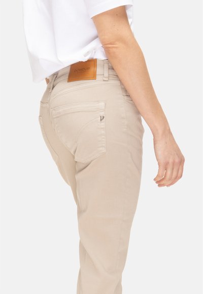 Pantaloni beige aderenti con dettaglio tasca posteriore e patch in pelle del marchio, indossati con una maglia bianca a maniche corte, vista laterale e posteriore.