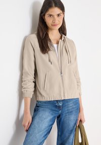 Veste zippée beige clair avec capuche, poignets et ourlet élastiques, deux poches latérales, et une texture lisse. Portée avec un jean bleu.