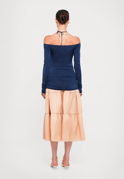 Jūras spēku zils off-shoulder tops, izgatavots no elastīga materiāla, apvienots ar gaiši persiku krāsas kārtainu svārku. Apģērbs ir aprīkots ar garām piedurknēm un saišu aizmugurējām detaļām.