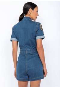 Jeansowy kombinezon z krótkimi rękawami, jasnoniebieski kolor, widoczne szwy, talia z paskiem oraz naszywkami na ramionach. Rękawy zakończone podwinięciem.