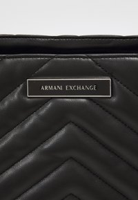 Must mustril pistetud nahk käekott, millel on chevron-mustrid ja hõbedane bränditud plaat, millel on kiri "ARMANI EXCHANGE."