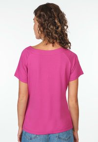 Fuchsia Kurzarmoberteil mit Bootsausschnitt. Hat eine glatte Textur und eine lockere Passform, von hinten gesehen mit sichtbaren lockigen Haaren.