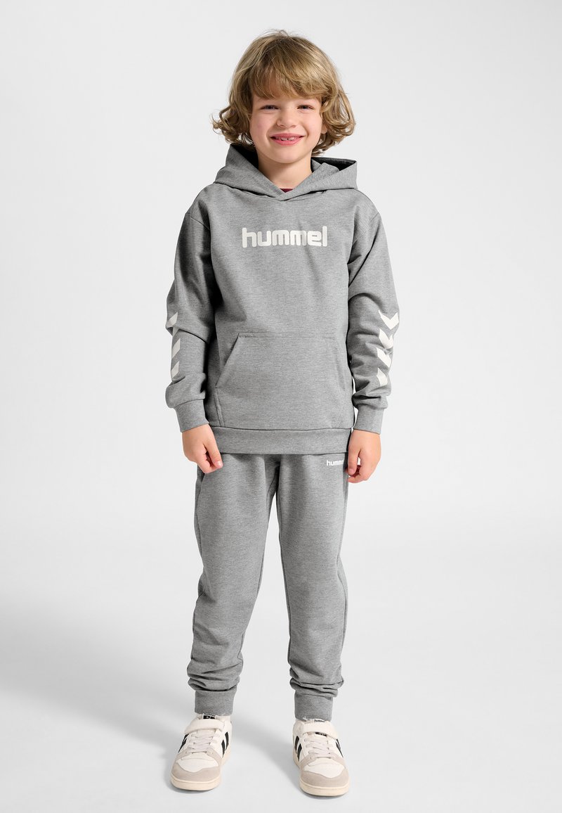Lille barn står og smiler, iført grå hummel hættetrøje og matchende joggingbukser med hvide sneakers mod en ensfarvet baggrund.