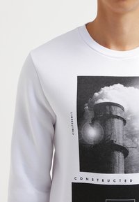 Weißes Sweatshirt mit rundem Hals und langen Ärmeln; es verfügt über einen grafischen Druck eines Kamins und Wolken in Schwarz und Grau.