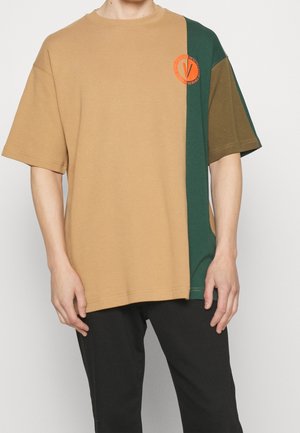 Mann trägt ein kurzärmeliges beige-grünes T-Shirt mit einem orangefarbenen runden Logo auf der linken Brust.