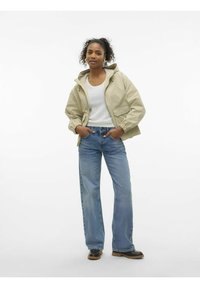 Beige Kapuzenjacke mit elastischen Bündchen, getragen über einem weißen Pullover. Kombiniert mit hellblauen weiteren Jeans und schwarzen Stiefeln.