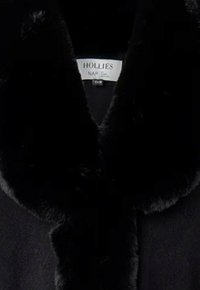 Svart kappa med en fuskpälskrage, slät textur och djup V-ringning. Etiketten säger "HOLLIES NAP."