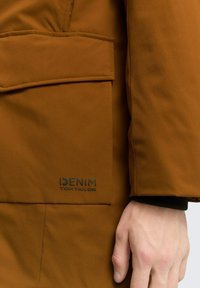Smeđi kaput od glatke tkanine, sa prednjim džepom i logotipom koji glasi "DENIM TOM TAILOR." Ruka s crnom manžetom je vidljiva.