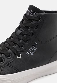 Mustad nahast kesk kõrguse sneaker, millel on tekstureeritud pind, mustad nöörid, hõbedased silmad ja valge GUESSi logo küljel.