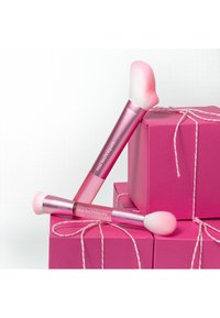 Twee roze make-up kwasten met witte haren die rusten op en leunen tegen roze cadeaudoosjes die zijn dichtgebonden met roze en wit touw.