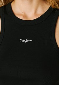 Prim-plan cu o persoană purtând un top negru fără mâneci, cu model reiat, cu logo-ul „Pepe Jeans” brodat alb la nivelul pieptului.