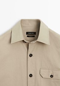 Beige bomulds skjorte med klassisk krave, store sorte knapper og en brystlomme med flap. Etiketten læser "Massimo Dutti."