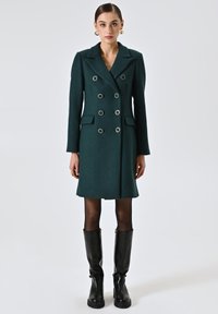 Bosgroene dubbelzijdige jas met een geweven patroon, grote metalen knopen, zijzakken en een getailleerde pasvorm, gecombineerd met kniehoge zwarte laarzen.
