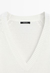 Hvid strikket V-hals sweater med ribbet krave og et sort mærke med brand og oprindelse inde i halsudskæringen.