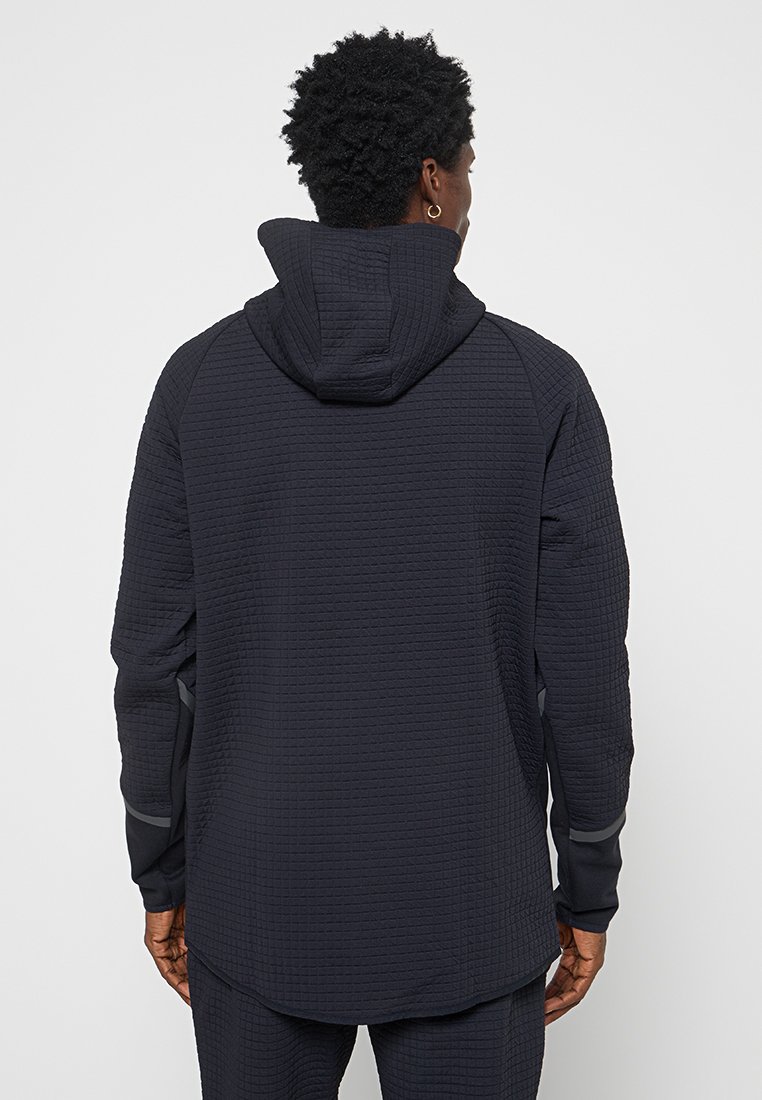 Giacca con cappuccio grigio scuro realizzata in un tessuto trapuntato e texture. Presenta maniche raglan e piccoli dettagli riflettenti.