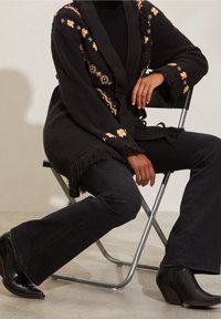 Black knit cardigan with floral embroidery, fringed edges. Paired with black flared jeans and black wedge ankle boots. Sitting on a chair.