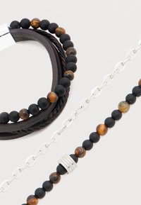 Bracelets en cuir tressé et perlé avec des perles noires et marron, ainsi qu'un bracelet à maillons en argent sur fond blanc.