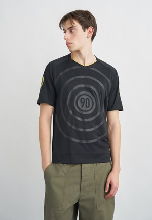 Jeune homme portant une chemise noire ornée de cercles concentriques et du chiffre 90, debout les mains dans les poches, regardant vers sa gauche.