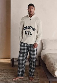 Lys beige kvart-zip sweatshirt med "BROOKLYN NYC" tekst. Sort og hvid ternet pyjamabukser. Tekstureret stof, afslappet pasform.