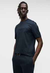 Μπλούζα μπλε ναυτικού με στρογγυλή λαιμόκοψη, κατασκευασμένη από απαλό ύφασμα, που διαθέτει ανάγλυφο λογότυπο Aston Martin στο στήθος, συνδυασμένη με σκουρόχρωμο, άνετης εφαρμογής παντελόνι.