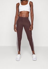Hnědé legíny s bílými bočními pruhy v kombinaci s bílým sportovním podprsenkou. Tkanina vypadá hladce, navržena pro aktivní nošení. Bílé tenisky dokončují vzhled.
