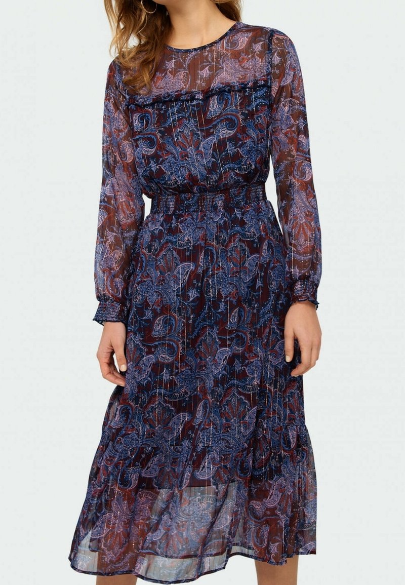 Žena v dlouhých rukávech, průhledných námořnických midi šatech s fialovým a červeným květinovým paisley vzorem a elastickým pasem.
