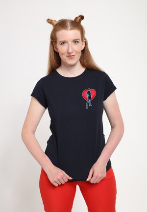 Navy bomulds t-shirt med et lille grafisk motiv af en kvinde i et hjerte-design. Modellen sætter den sammen med klare røde bukser. Korte ærmer.