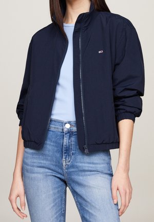 Kvinde iført navyblå bomberjakke med lynlås over lyseblå skjorte og lyseblå jeans, stående foran en ensfarvet baggrund.