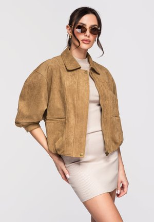 Femme portant une veste en daim marron sur une robe côtelée beige, accessoirisée de lunettes de soleil teintées marron et de boucles d'oreilles en forme d'anneau doré, posant avec la main sur la hanche.