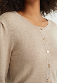 Kvinna som bär en beige stickad cardigan med runda guldknappar och rund halsringning, sedd från halsen till midjan.