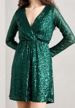 Femme portant une robe verte à sequins, à manches longues et col en V profond, avec une taille cintrée, debout les mains sur les hanches devant un fond uni.